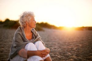 Older woman with short white hair sitting on a beach at sunset, smiling into the distance for health and wellness IGNITE Peak Performance coaching service