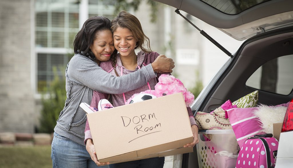 Mother hugging daughter behind open car, daughter is holding a box labeled Dorm