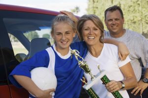 Man, mother, and their daughter standing leaning against a car outside. Daughter is an athlete wearing a jersey and holding a ball with her arm around mom, who is holding a trophy. Dad is standing behind mom. All three smiling at the camera for parent workshop IGNITE Peak Performance elite coaching service