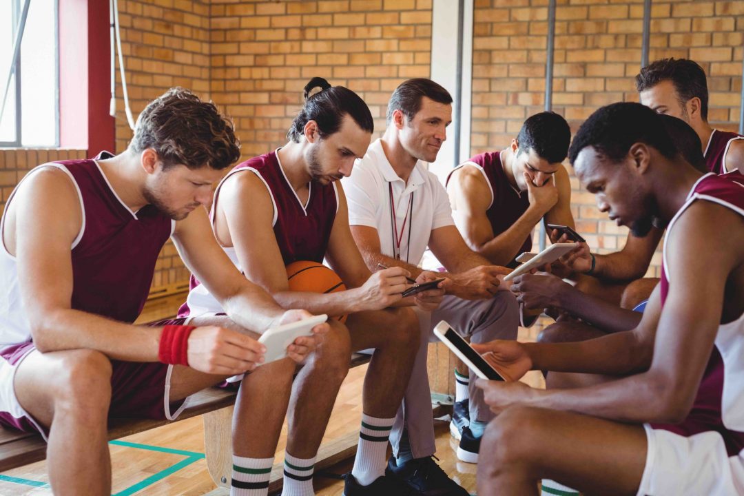 Basketball sport athlete team in maroon jerseys sitting together with coach or trainer, studying materials on benches on the court. Indicates team and group training offered by international coach at IGNITE Peak Performance elite coaching services