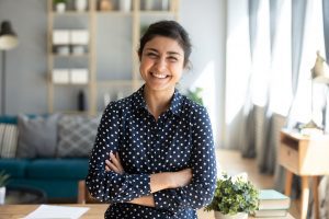 Professional woman standing in living room in a navy shirt, crossing her arms and smiling at the camera for personal and life IGNITE Peak Performance coaching service