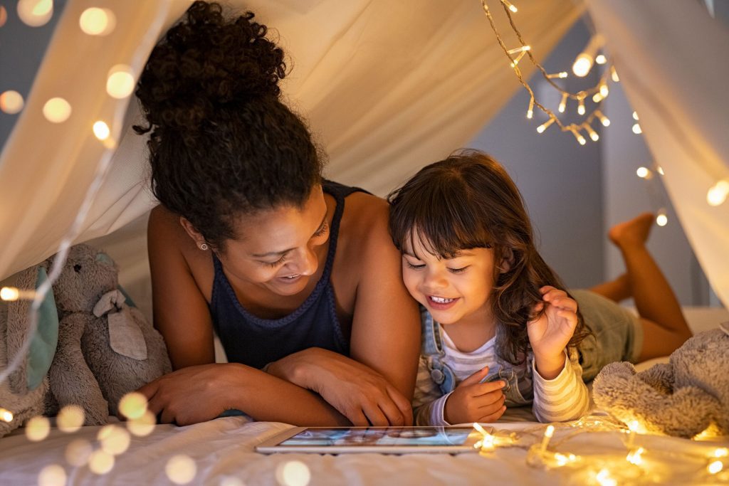 Mom and daughter smiling and reading a book together in a blanket fort with lights and stuffed animals for women and mothers IGNITE Peak Performance coaching service