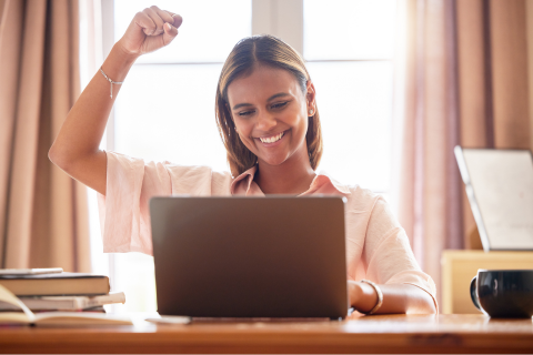 Image of smiling young woman at laptop and celebrating with her arm in the air