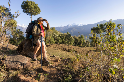 Image of older woman leaning on rock in a field and looking through binoculars while holding a hiking pole